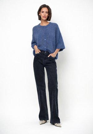 Vrouw staand met handen in de zakken, draagend een blauwe, pluizige trui met knopen, zwarte hooggetailleerde jeans en schoenen met een patroon en puntige neus.