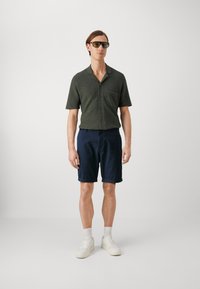 Camicia verde scuro a maniche corte con tasca frontale, abbinata a pantaloni corti blu e sneakers bianche. Gli accessori includono occhiali da sole. Design semplice.