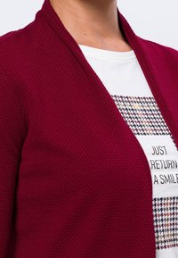 Burgunderfarbener Cardigan mit strukturiertem Strickdesign, offenem Front und ohne Verschlüsse, über einem weißen T-Shirt mit grafikdruck im Hahnentrittmuster.