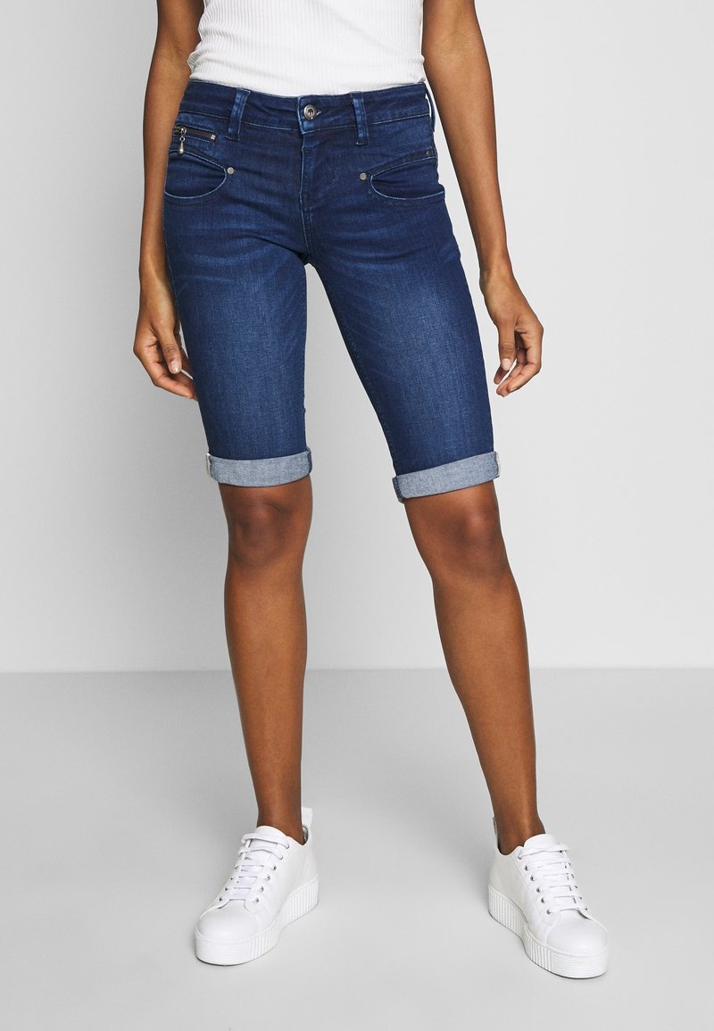 Person i uppvikta mörkblå denimshorts och vita sneakers, som står mot en enkel ljusgrå bakgrund.