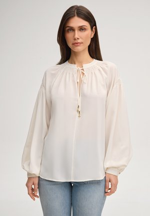 Kobieta o prostych ciemnych włosach, ubrana w luźną białą bluzkę z marszczonym dekoltem i długimi bufkowymi rękawami, w połączeniu z jasnoniebieskimi dżinsami.