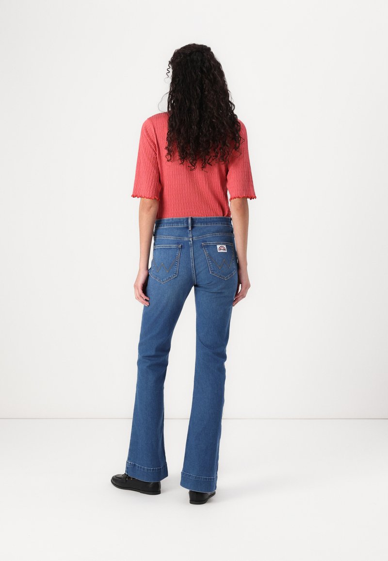 Jeans a zampa d'elefante in denim blu con vita alta, leggermente sbiaditi e tasche posteriori ricamate. Abbinati a un top corallo a costine e scarpe nere.