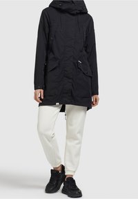 Svart parka-jacka med huva, som har framfickor, en lång passform och dragskoaccent, bärs över ljusa joggers och svarta sneakers.