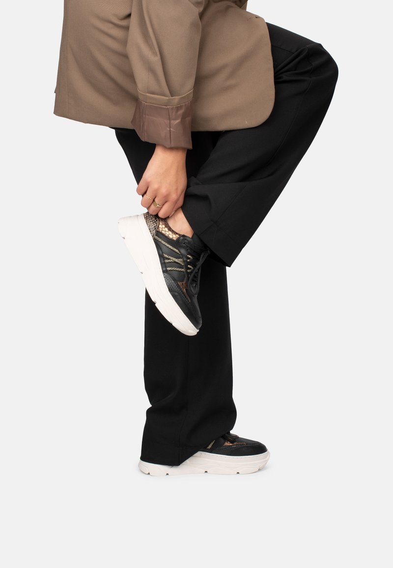 Black athletic shoes with a textured mesh upper and white sole, featuring golden accents and a lace-up design. Worn with black pants.