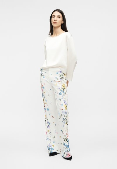 Γυναίκα με μακριά σκούρα μαλλιά, φορώντας λευκό πουκάμισο με μακριά μανίκια και λευκή παντελόνα με φαρδιά μπατζάκια διακοσμημένη με πολύχρωμο ανθισμένο μοτίβο και μαύρα παπούτσια.