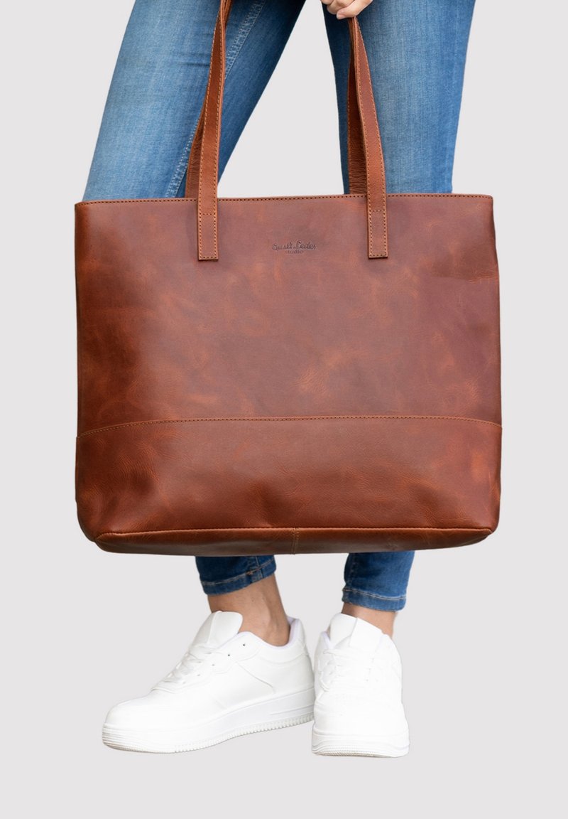 Bruine leren tote bag met twee lange handvatten, een gladde textuur en minimale stikdetails, vastgehouden voor een been gekleed in blauwe jeans.