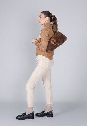 Bruine furrugzak met leren accenten, beige trui, crèmekleurige broek en zwarte loafers met sokken. Model poseert zijwaarts tegen een grijze achtergrond.