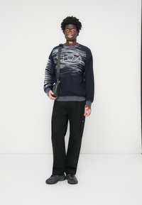 Marineblå sweatjakke med grå bølge mønster, ribbede mansjetter, kombinert med svarte cargo-bukser og svarte sneakers. Modellen holder en liten svart veske.