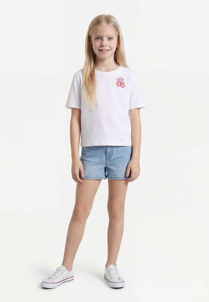 Mlada deklica z dolgimi blond lasmi, oblečena v belo majico z motivom češenj, svetlo modre jeans kratke hlače in bele superge na belem ozadju.