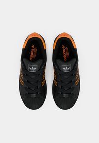 Sapatilhas Adidas pretas com detalhes em laranja, três riscas e ponta de borracha. O tecido texturizado e os atacadores pretos planos oferecem suporte e estilo.