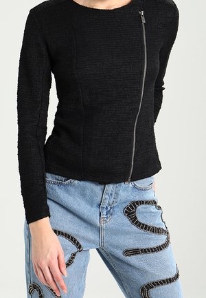 Woman wearing a fitted black textured long-sleeve top with an asymmetrical front zipper and light blue jeans with black beaded snake designs.