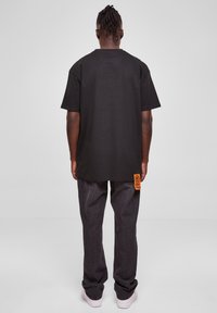Black short-sleeve T-shirt with a relaxed fit, paired with dark denim jeans. Notable orange branding tag on the left side seam.
