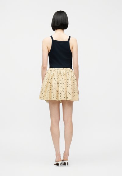 Γυναίκα με κοντά μαύρα μαλλιά στέκεται γυρίζοντας την πλάτη, φοράει μαύρο αμάνικο τοπ, μπεζ φούστα με πουά και λευκά σανδάλια με τακούνι.