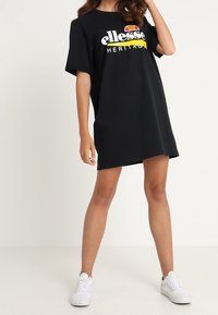 Schwarzes, übergroßes T-Shirt-Kleid mit kurzen Ärmeln, das ein buntes, gedrucktes Logo auf der Brust trägt. Kombiniert mit weißen Sneakers.