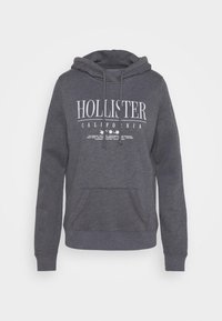 Grå bomullshuvtröja med en framficka, dragsko i huvan och vit tryckt text "HOLLISTER CALIFORNIA" på framsidan.