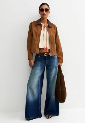 Kobieta w brązowej kurtce na kremowej bluzce z wiązaniem, szerokie niebieskie jeansy, okulary przeciwsłoneczne, kolczyki koła, trzymająca brązową torbę przy boku.