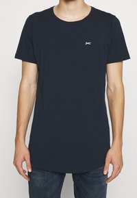 Námorně modré bavlněné tričko s kulatým výstřihem, krátkými rukávy a zakřiveným lemem. Na hrudi se nachází malé bílé logo nůžek.