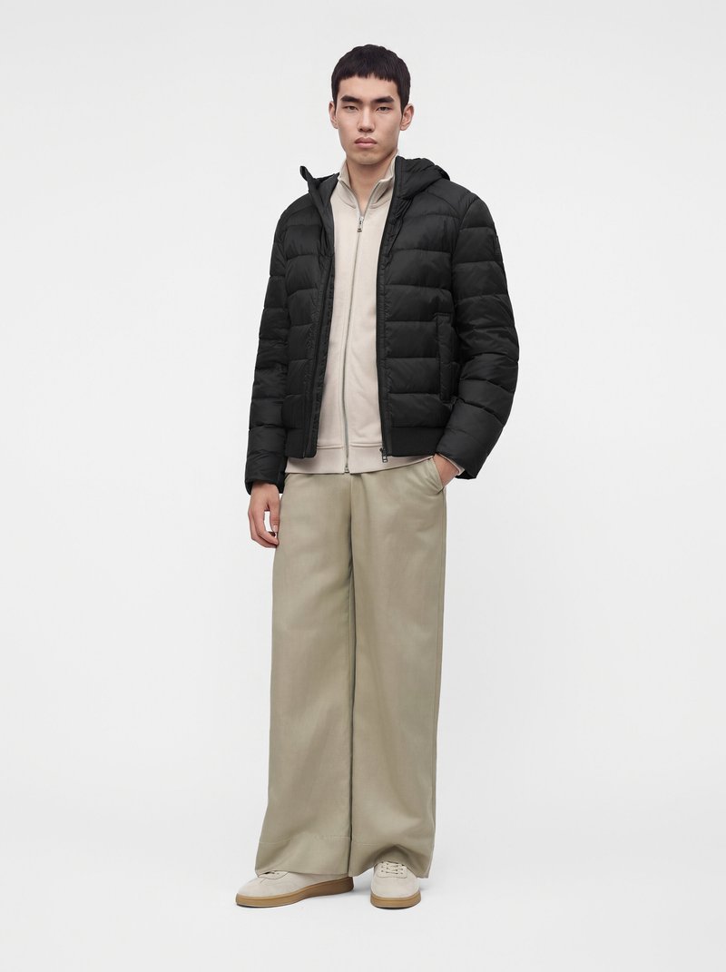 Jeune homme portant une doudoune noire sur un haut beige zippé, un pantalon large beige et des baskets claires, debout devant un fond uni.