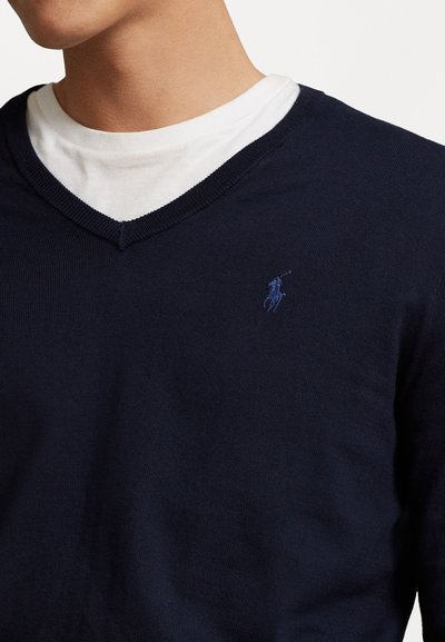Marine gebreide V-hals trui, gelaagd over een witte ronde hals shirt, met een subtiel geborduurd logo op de linkerborst. Glad textuur.