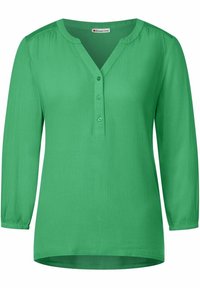 Blouse verte à manches longues en tissu texturé. Présente un décolleté en V, une patte de boutonnage à cinq boutons et des poignets froncés. Ourlet arrondi.