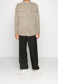 Beige fløjlsjakke med knaplukning på brystlommer, kombineret med løstsiddende sorte bukser og hvide sneakers.