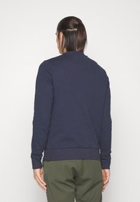 Granatowa bluza z długimi rękawami, ściągaczami na mankietach i ściągaczem u dołu. Gładka faktura, okrągły dekolt, widok od tyłu.