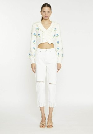 Mujer de pie, de frente, lleva un jersey corto blanco con bordado de flores azules, vaqueros blancos rotos y sandalias de tacón beige.