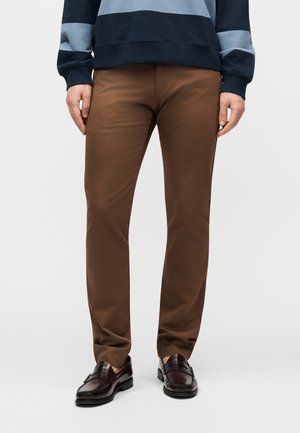 Homme portant un pantalon brun slim et des mocassins en cuir brun foncé, debout avec les mains détendues le long du corps sur un fond uni.