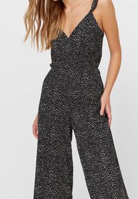 Černý overal s výstřihem do V, širokými nohavicemi a tenkými ramínky. Disponuje bílým tečkovaným vzorem. Vyroben z měkké, lehké látky.