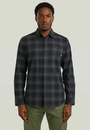 Mand iført en sort og gråternet skjorte med knapper og brystlomme, kombineret med olivengrønne bukser, stående foran en ensfarvet baggrund.