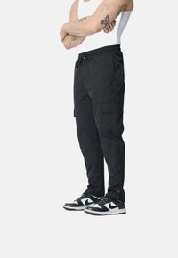 Pantalons cargo noirs avec une taille élastique, cordon de serrage et plusieurs poches latérales. Portés avec des baskets noir et blanc.