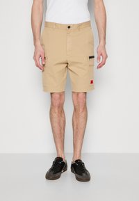 Pantalones cortos cargo beige con dos bolsillos laterales y un bolsillo con cremallera. Hechos de algodón con una textura suave. Incluye un botón negro y un detalle de etiqueta roja.
