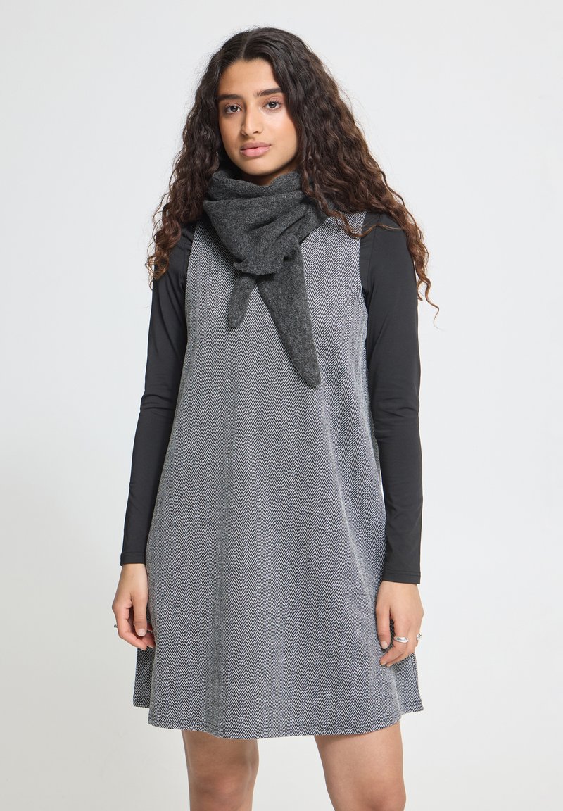 Jeune femme aux longs cheveux bouclés portant une robe gris chevron par-dessus un t-shirt noir à manches longues et une écharpe gris foncé sur un fond uni.