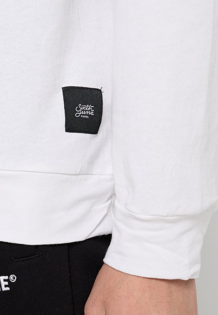 Witte blouse met lange mouwen, voorzien van een zwart stoffen label op de mouw, met een zachte textuur en een ontspannen pasvorm. Zwarte broek gedeeltelijk zichtbaar.