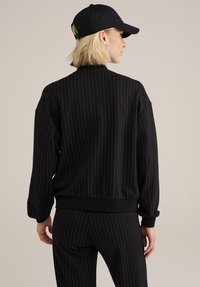 Črno bomber jakna z drobnimi črtami, mehka na otip, z rebrastimi manšetami in robom. Nosite jo z ujemajočimi hlačami in črno kapo.
