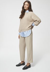 Beżowy sweter w robótkach włożony na jasnoniebieską koszulę z kołnierzykiem, beżowe szerokie spodnie i czarne mokasyny na wsuwkę z metalowym akcentem.