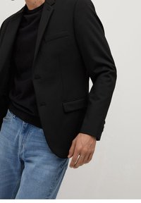 Musta räätälöity bleiseri, joka on valmistettu sileästä kankaasta, jossa on yksi rintatasku ja halkiotaskut, yhdistettynä slim fit mustaan villapaitaan ja vaaleansinisiin farkkuihin.