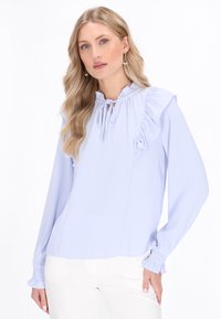 Lyseblå bluse med flæse detaljer ved skuldrene, snøre ved halsen og rynkede manchetter. Glat stof med en tætsiddende pasform.