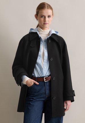 Zwarte wolcoat met brede revers, grote knopen en zijzakken. Daaronder een lichtblauw gestreept overhemd, laag over een witte turtleneck.