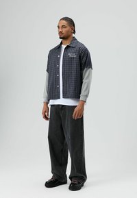 Zwart-grijs geruit shirt met korte mouwen, gedragen over een grijs shirt met lange mouwen, gecombineerd met donkere, losse jeans en zwarte schoenen.