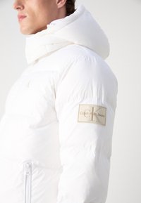 Vit puffjacka med en rynkad textur, med hög krage och Calvin Klein Jeans logotyp i beige på vänster ärm.