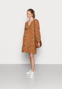 Vestido de padrão floral na cor laranja enferrujada, com decote em V, mangaslongas e bainha evasê, combinado com ténis brancos e uma bolsa de ombro castanha.