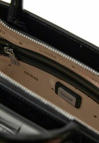 Sac à main synthétique noir avec fermeture zippée, intérieur beige imprimé du logo "GUESS" et étiquette en métal argenté.