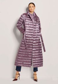 Lange, lichtpaarse gewatteerde jas met een quilted ontwerp, hoge kraag, strik taille en zijzakken, gecombineerd met donkere jeans en hoge hakken.