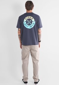 T-shirt en coton gris avec un graphique circulaire bleu et jaune au dos, accompagnée d'un pantalon beige clair et de baskets noires.