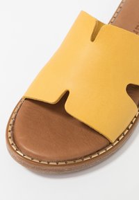 Öppen tå sandal med ovandel i gult läder och brun vadderad sula, med dekorativ stygn runt sula kanten.