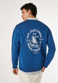 Uomo che indossa un felpa blu con una grafica di squalo e il testo Pepe Jeans London, sovrapposta a una camicia a righe con colletto, pantaloni bianchi.