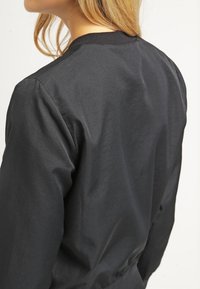 Czarna kurtka bomberka o gładkiej fakturze, z prążkowanym kołnierzem i dopasowanymi rękawami. Widok z tyłu pokazuje minimalistyczny design bez ozdób.