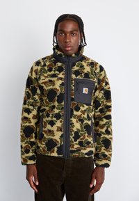 Veste en polaire avec un motif camouflage vert et marron, dotée d'une fermeture éclair noire et d'une poche avec logo. Texture douce et coupe décontractée.