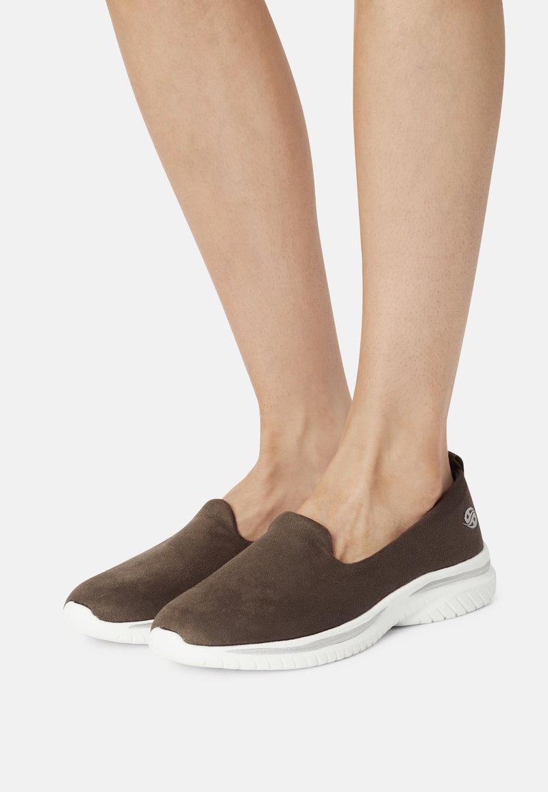 Chaussures brunes à enfiler sur des jambes nues, mettant en valeur une semelle blanche rembourrée et un matériau doux. Pieds positionnés pour une vue de côté.